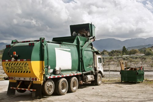 A large green waste collection truck with a yellow caution stripe and reflective markings is parked on a dirt and gravel surface, facing towards the right side of the image. The truck's rear hopper is open, indicating it is in the process of collecting or about to collect rubbish. To the right of the truck, there is a green skip bin filled with waste materials, positioned on the ground next to the vehicle. The environment appears to be an outdoor industrial or rubbish disposal area, with a backdrop of low mountains and a partly cloudy sky, suggesting daytime conditions. The scene lacks nearby structures but includes visible fencing in the background and general signs of waste management activity consistent with local rubbish removal services, such as those offered by Maida Vale House Clearance in London postcode areas. The overall setting emphasizes efficient waste collection and disposal, typical of professional rubbish management operations in the region.